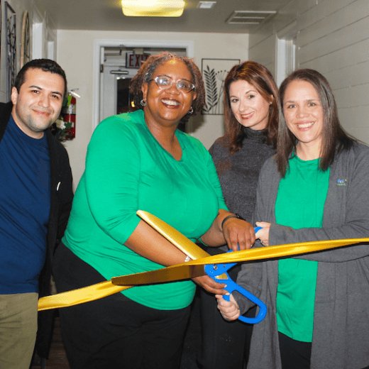 Group of colleagues smiling and holding large scissors to cut a yellow ribbon.