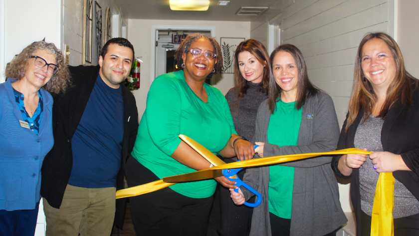 Group of colleagues smiling and holding large scissors to cut a yellow ribbon.