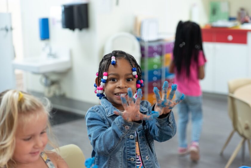 Child smiling and holding up hands covered in blue paint.