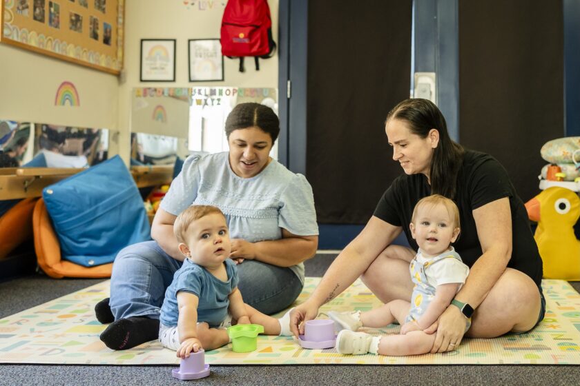 Two woman and two babies sitting on a rug playing.