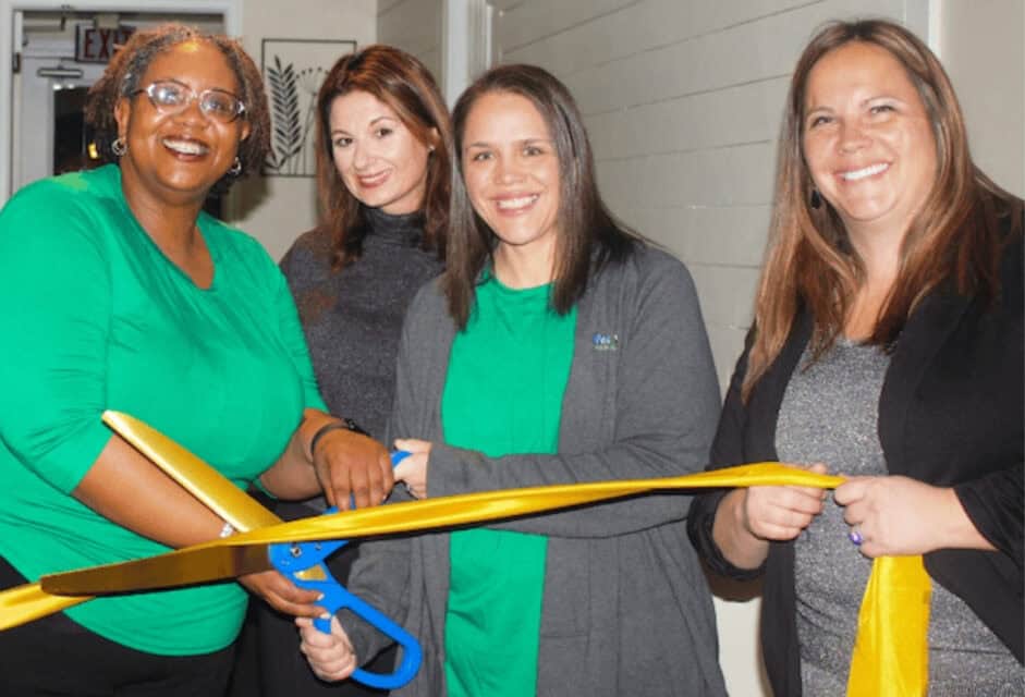 Group of colleagues smiling and holding large scissors to cut a yellow ribbon.